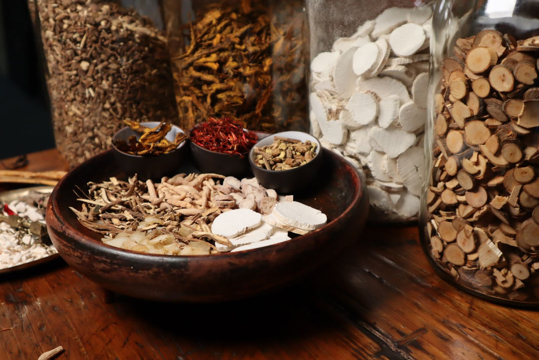 A wooden bowl of mixed dried herbs and roots beside glass storage jars