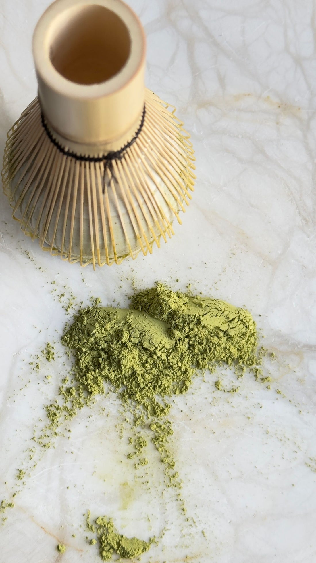 Bright green matcha powder beside a bamboo whisk on a pale surface