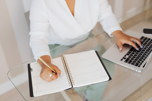 Person writing in a notebook while using a laptop at a desk, focused work